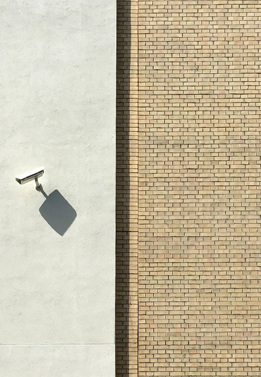 White surveillance camera on a pale wall, its shadow falling diagonally, next to a strip of yellow brick