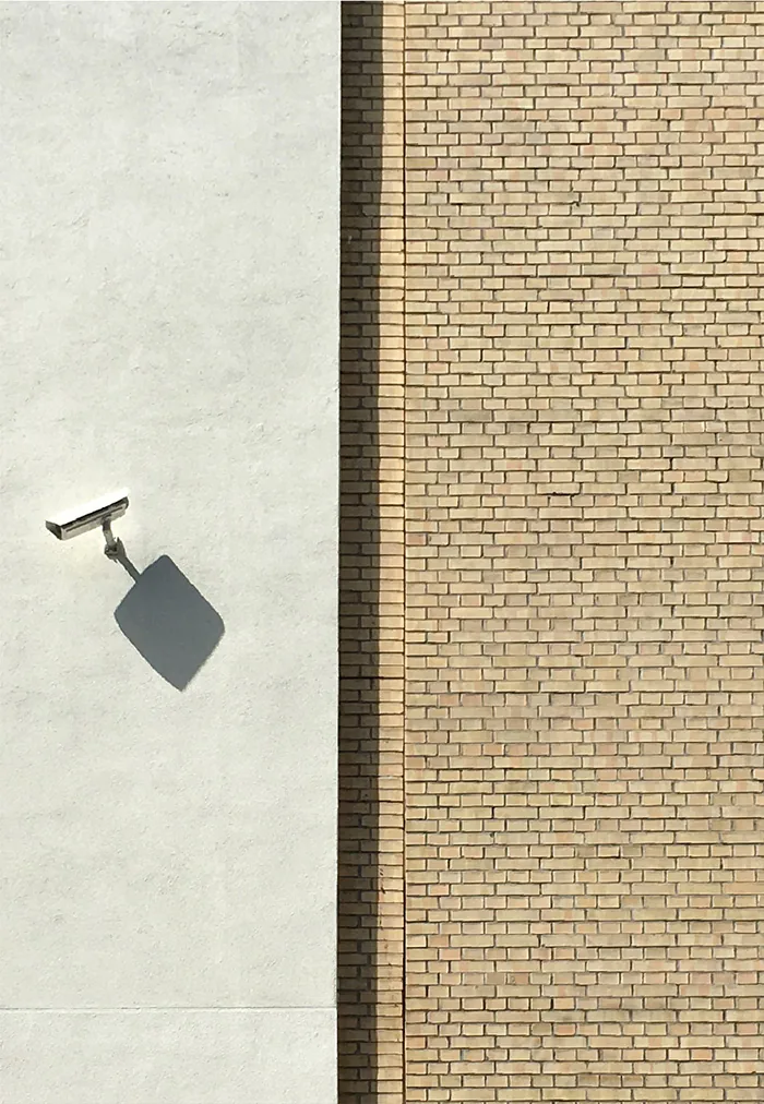 White surveillance camera on a pale wall, its shadow falling diagonally, next to a strip of yellow brick
