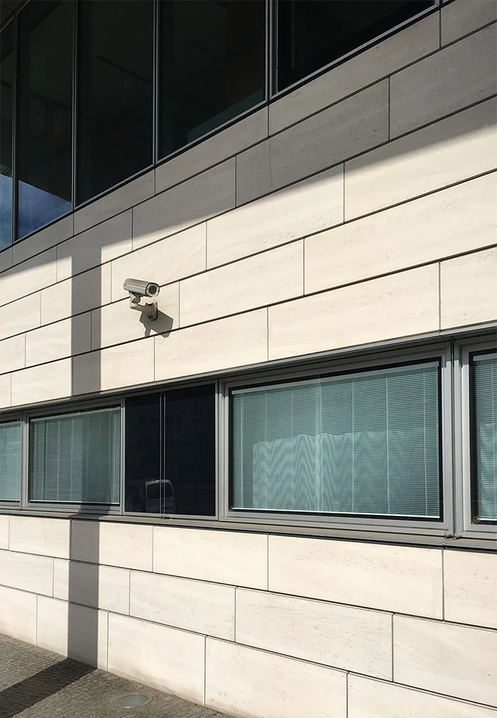 Camera on a light stone facade between bands of dark windows, in raking sunlight