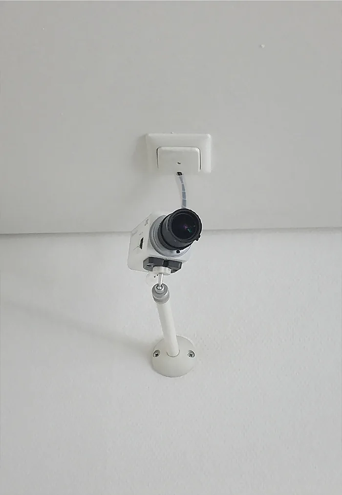White camera on a bracket below a light switch on a plain white wall
