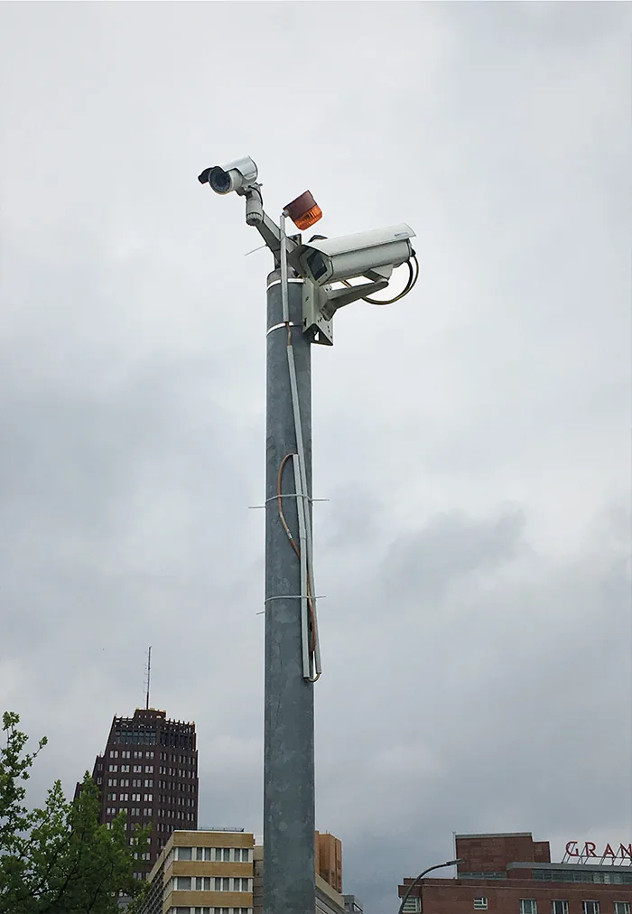 Tall pole with several cameras against a grey sky, city buildings below