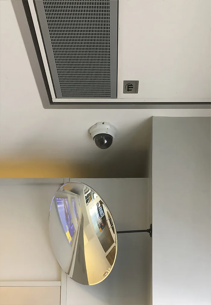Dome camera on a ceiling above a convex safety mirror in a white hallway