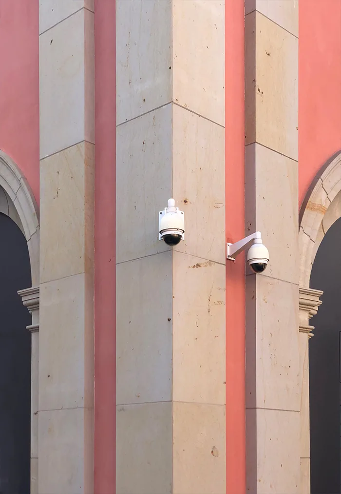 Two dome cameras on pale stone pillars in front of a pink wall
